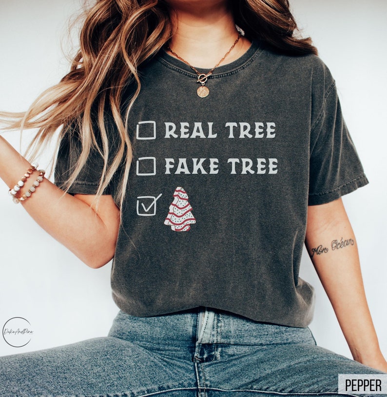 May include: Dark gray t-shirt with the text "REAL TREE" and "FAKE TREE" in white. A Christmas tree graphic is checked, indicating a preference for a real tree. The shirt is a comfortable, casual style.