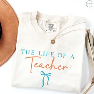 Puede incluir: Camiseta color crema con el texto "THE LIFE OF A Teacher" en turquesa y naranja, con un lazo turquesa. También se ven un collar dorado con un colgante y un sombrero marrón.