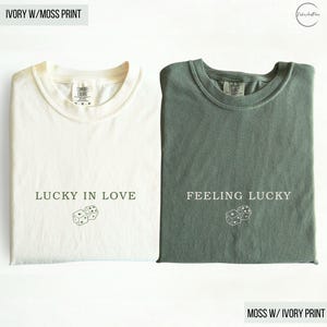 May include: Two folded t-shirts, one ivory and one moss green. The ivory shirt has the text "LUCKY IN LOVE" and dice graphic. The moss green shirt has the text "FEELING LUCKY" and a dice graphic. Both shirts are Comfort Colors brand.