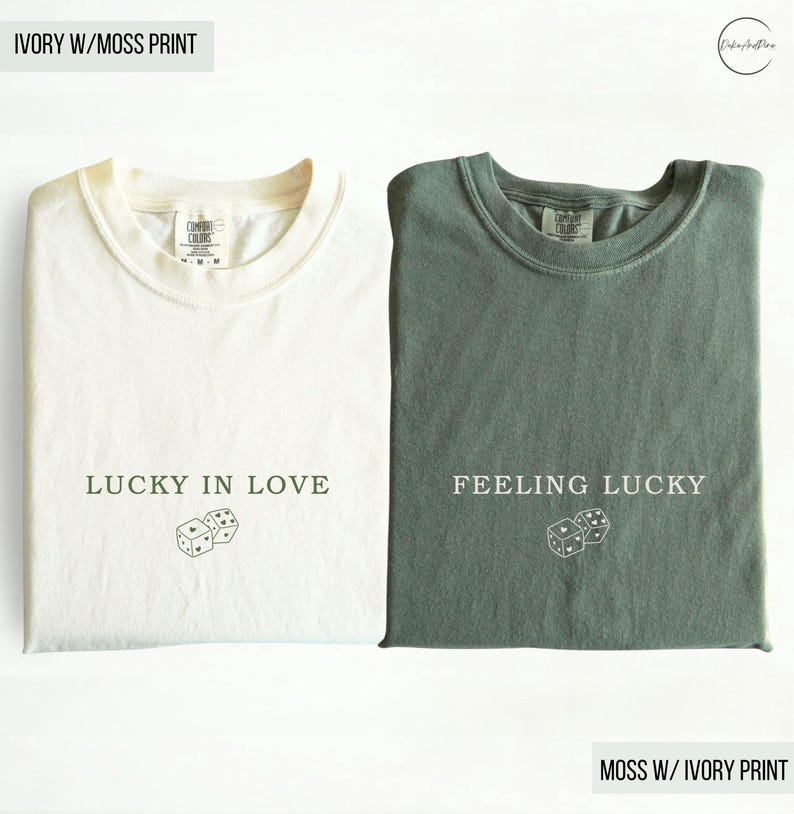 May include: Two folded t-shirts, one ivory and one moss green. The ivory shirt has the text "LUCKY IN LOVE" and dice graphic. The moss green shirt has the text "FEELING LUCKY" and a dice graphic. Both shirts are Comfort Colors brand.
