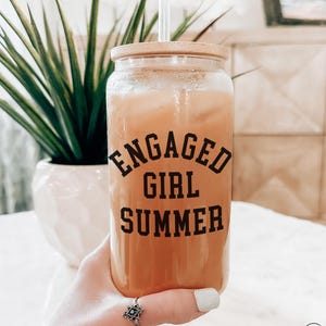 May include: A clear glass tumbler with a bamboo lid and straw, filled with an orange beverage. The words "ENGAGED GIRL SUMMER" are printed in black. The tumbler is held by a person wearing a ring.