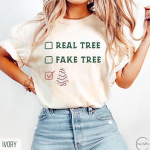May include: Ivory-colored t-shirt with the text "REAL TREE" and "FAKE TREE" in green, with a checkmark next to a decorated Christmas tree graphic. The shirt is paired with blue jeans.