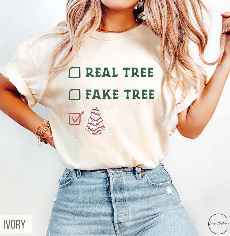 May include: Ivory-colored t-shirt with the text "REAL TREE" and "FAKE TREE" in green, with a checkmark next to a decorated Christmas tree graphic. The shirt is paired with blue jeans.