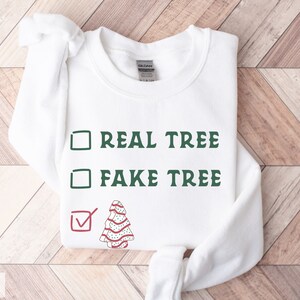 May include: White sweatshirt with the text "REAL TREE" and "FAKE TREE" in green, with a checkmark next to a decorated Christmas tree graphic. The sweatshirt is a Gildan brand.