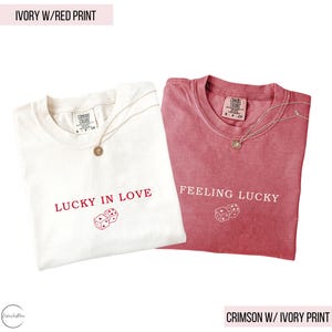May include: Two t-shirts, one ivory and one crimson, are displayed. The ivory shirt has red text that reads "LUCKY IN LOVE" with dice graphics. The crimson shirt has ivory text that reads "FEELING LUCKY" with dice graphics. Both shirts have a necklace.