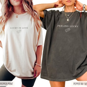 May include: Two t-shirts, one ivory and one pepper, each with a graphic print. The ivory shirt has the text "LUCKY IN LOVE" above a dice graphic. The pepper shirt has the text "FEELING LUCKY" above a dice graphic.