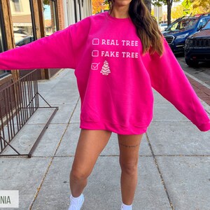 May include: A bright pink sweatshirt with the text "REAL TREE" and "FAKE TREE" in white. A small Christmas tree graphic is below the text. The sweatshirt is a crew neck style.