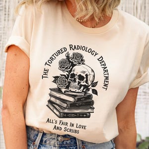 May include: A beige t-shirt with a black and white graphic design. The graphic features a skull with roses and books. The text reads "The Tortured Radiology Department" and "All's Fair in Love and Scrubs".