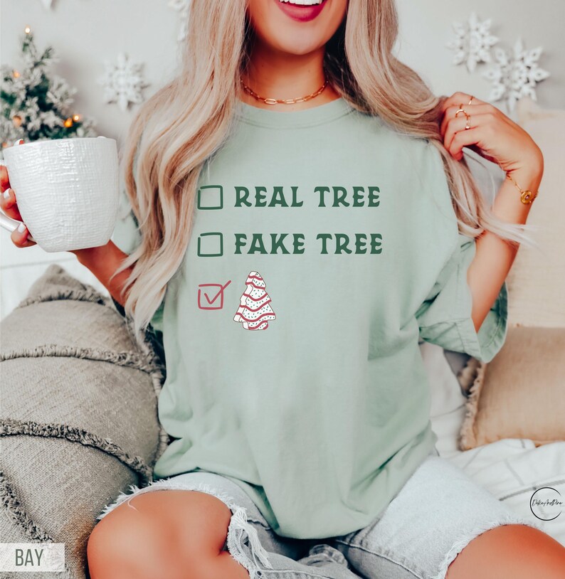 May include: A light green t-shirt with the text "REAL TREE" and "FAKE TREE" in dark green. A red checkmark is next to a drawing of a decorated Christmas tree. The shirt is a casual style.