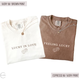 May include: Two t-shirts, one ivory and one espresso, with the text "LUCKY IN LOVE" and "FEELING LUCKY" printed on them. Each shirt has a dice graphic and a gold necklace. The text "IVORY W/ BROWN PRINT" and "ESPRESSO W/ IVORY PRINT" are also visible.