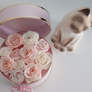 May include: A pink gift box with a gold trim lid is open, revealing a bouquet of delicate pink and white roses. The box is sitting on a white surface next to a ceramic cat figurine.