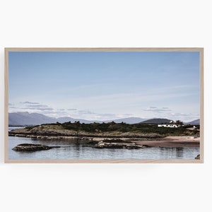 Pode incluir: Uma fotografia de paisagem que mostra uma pequena casa branca em uma península rochosa com uma massa de água em primeiro plano e uma cordilheira ao fundo. O céu é azul claro com algumas nuvens.