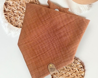 May include: A rust-colored dog bandana with a textured, crinkled appearance. The bandana is folded and has a small leather tag with the text "Little Paw". The bandana is displayed on a white surface with woven placemats.