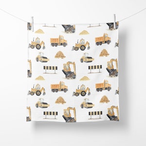 May include: A white muslin baby swaddle with a repeating pattern of yellow construction vehicles, including dump trucks, excavators, and road rollers. The vehicles are surrounded by small piles of sand.