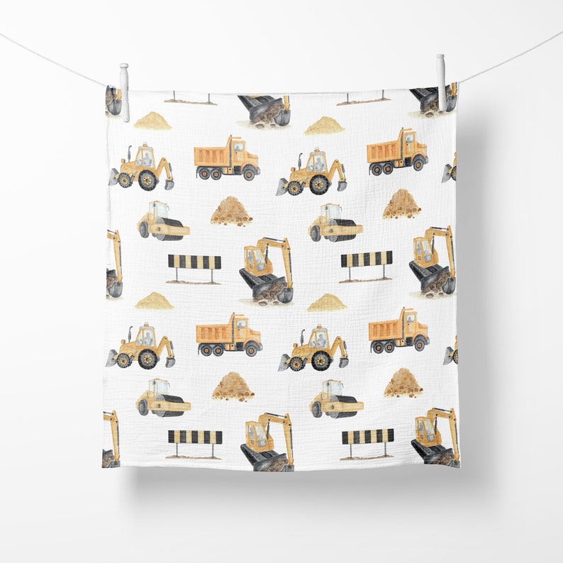May include: A white muslin baby swaddle with a repeating pattern of yellow construction vehicles, including dump trucks, excavators, and road rollers. The vehicles are surrounded by small piles of sand.