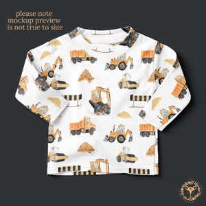 May include: White long-sleeved shirt with a repeating pattern of yellow construction vehicles, including excavators, dump trucks, and road rollers. The vehicles are painted in a watercolor style on a white background.