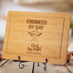 May include: A rectangular bamboo cutting board with a carved border, resting on a black metal stand. The board features the text "ENGINEER BY DAY" above a chef's hat and crossed knives, with "Chef by night" and space for a name below.