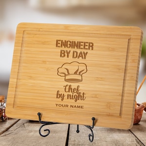 May include: A rectangular bamboo cutting board with a carved border, resting on a black metal stand. The text "ENGINEER BY DAY" and "Chef by night" with a chef's hat graphic are engraved on the board. Space is provided for a name.