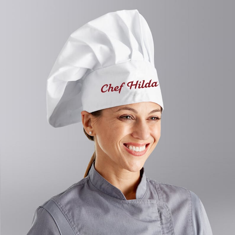 Puede incluir: Sombrero de chef blanco con el texto "Chef Hilda" bordado en rojo en la parte delantera.