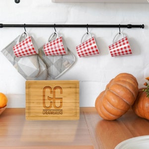 Puede incluir: Escena de cocina con una tabla de cortar de bamb&uacute; grabada con las iniciales "CG" y el texto "CREATIVEGEEKS". Tazas a cuadros rojos y blancos cuelgan de una barra negra. Tambi&eacute;n se ven dos calabazas y una cesta de frutas.