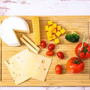 Puede incluir: Una tabla de cortar de madera con una variedad de alimentos. Incluye una rueda de queso brie, cubos de queso cheddar, queso en rodajas, palitos de pan, tomates cherry, br&oacute;coli y dos tomates grandes. El fondo es blanco con vetas de madera.