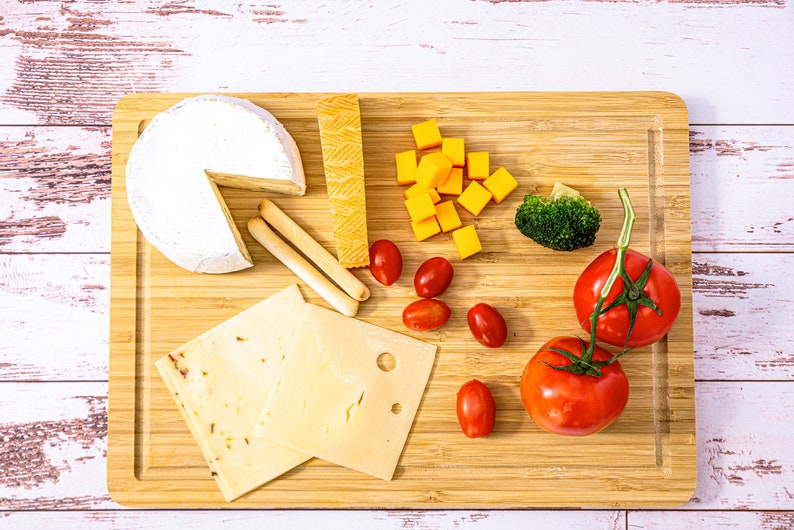 Puede incluir: Una tabla de cortar de madera con una variedad de alimentos. Incluye una rueda de queso brie, cubos de queso cheddar, queso en rodajas, palitos de pan, tomates cherry, br&oacute;coli y dos tomates grandes. El fondo es blanco con vetas de madera.