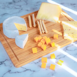 Puede incluir: Una tabla de cortar de madera muestra una variedad de quesos, incluyendo una rueda de brie, cheddar en rodajas y queso en cubos. Palitos de pan est&aacute;n dispuestos entre los quesos. Los quesos est&aacute;n dispuestos en una tabla de cortar de madera.