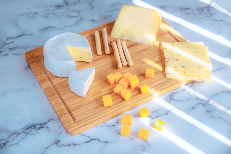 Puede incluir: Una tabla de cortar de madera muestra una variedad de quesos, incluyendo una rueda de brie, cheddar en rodajas y queso en cubos. Palitos de pan est&aacute;n dispuestos entre los quesos. Los quesos est&aacute;n dispuestos en una tabla de cortar de madera.