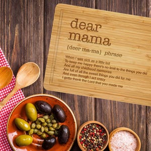 Puede incluir: Una tabla de cortar rectangular de bamb&uacute; con las palabras "dear mama" grabadas y un poema. La imagen tambi&eacute;n incluye un plato de aceitunas y alcaparras, cucharas de madera y peque&ntilde;os cuencos de especias y sal.