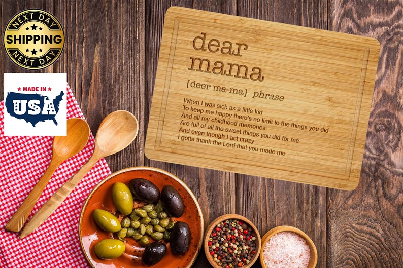 Puede incluir: Una tabla de cortar rectangular de bamb&uacute; con las palabras "dear mama" grabadas y un poema. La imagen tambi&eacute;n incluye un plato de aceitunas y alcaparras, cucharas de madera y peque&ntilde;os cuencos de especias y sal.
