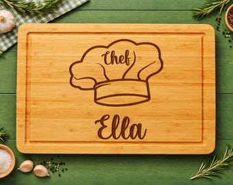 May include: A rectangular bamboo cutting board featuring a laser-engraved chef hat icon centered at the top with the word chef written inside. Below the hat, the name Ella is engraved in a cursive script. The board has a grooved border.