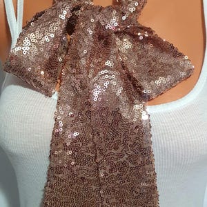 May include: A rose gold sequin scarf tied in a bow. The scarf is made of small, shiny sequins and is draped over a white tank top. The scarf's bow is centered, with the ends hanging down.