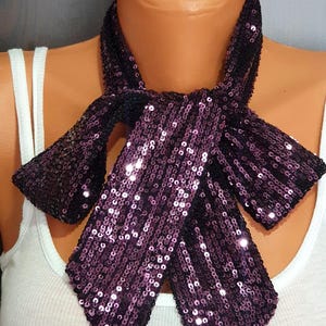 May include: A sequined bow tie in shades of purple and black, displayed on a mannequin. The bow tie is made of small, reflective sequins that catch the light. The accessory is worn over a white tank top.