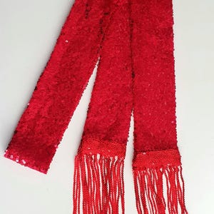 Peut inclure: Une écharpe rouge vif à paillettes avec de longues franges. L'écharpe est faite d'un matériau brillant et réfléchissant et est conçue pour être portée autour du cou ou de la taille. La frange ajoute du mouvement et du style.