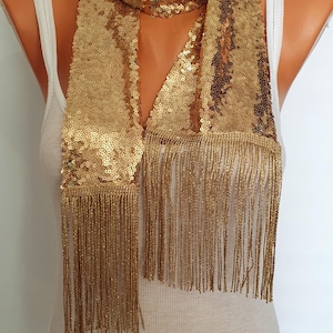 May include: A gold sequined scarf with long fringe. The scarf is draped around a mannequin's neck.