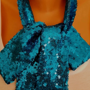Peut inclure: Une écharpe à paillettes turquoise est drapée autour du cou d'un mannequin. L'écharpe est faite de petites paillettes rondes et brillantes. L'écharpe est nouée en nœud au cou et a deux extrémités longues et pointues. Le mannequin porte un débardeur blanc.