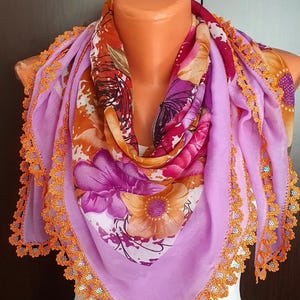 May include: A purple and orange floral scarf with a crocheted orange trim. The scarf is draped over a white mannequin.