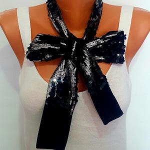Peut inclure: Un nœud papillon noir à paillettes avec des accents argentés, présenté sur un mannequin. Le nœud papillon est fait d'une matière semblable à du velours et est conçu pour être porté autour du cou. Le fond est blanc uni.