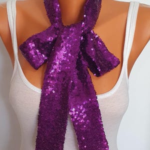 May include: A vibrant purple sequin scarf, tied in a bow. The scarf is draped around a mannequin's neck, showcasing its shimmering texture and design. The background is plain white.