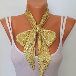 Peut inclure: Écharpe cravate à nœud papillon dorée à paillettes présentée sur un mannequin. L'écharpe est faite de petites paillettes dorées brillantes et est nouée en un grand nœud à l'avant. L'écharpe est autour du cou d'un mannequin portant un débardeur blanc.