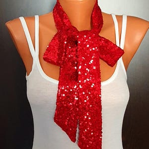 May include: A bright red sequined scarf tied in a knot around the neck, displayed on a white tank top. The scarf is covered in small, reflective red sequins that create a shimmering texture across the entire surface of the long, narrow fabric.