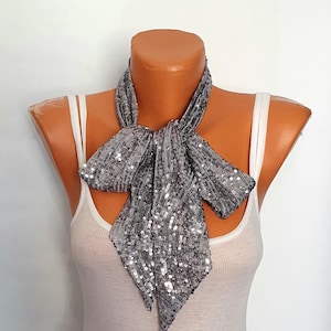 May include: A silver sequin scarf tied in a bow. The scarf is draped around a mannequin's neck, over a white tank top. The sequins reflect light, creating a shimmering effect. The background is plain white.