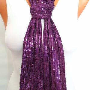 May include: A long, narrow scarf completely covered in small, reflective purple sequins. The scarf is tied in a simple knot at the neck, with two long, rectangular ends hanging down vertically. The fabric is dense with shimmering, circular sequins.