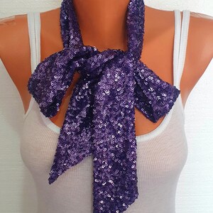 May include: A long, narrow scarf completely covered in small, reflective purple sequins. The scarf is tied in a simple knot at the center, with two ends hanging down. It is displayed on a mannequin wearing a plain white ribbed tank top.