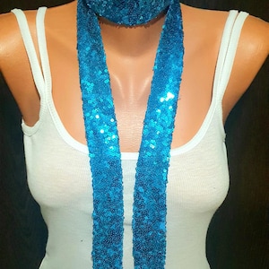 Op de afbeelding: Een felblauwe sjaal met lovertjes en een bijpassende halsband. De lange, smalle sjaal is bedekt met kleine, reflecterende blauwe lovertjes die een glinsterende textuur creëren. Het accessoire wordt getoond op een etalagepop met een wit hemdje.