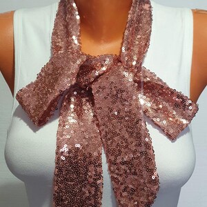 May include: A rose gold sequined scarf tied in a bow. The scarf is made of small, shimmering sequins and is a festive accessory.