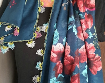 Pode incluir: Um lenço verde-azulado escuro com uma grande estampa floral de pétalas vermelho-profundo e azul-claro. As bordas do lenço são finalizadas com um intrincado acabamento de renda de agulha multicolorida em tons de rosa, amarelo, branco e azul-claro.