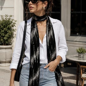 May include: A long black scarf featuring a vertical pattern of shimmering silver sequins, worn wrapped around the neck. The scarf is paired with a crisp white button-down shirt, light blue denim jeans, and a black shoulder bag with a thin strap.