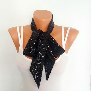 May include: A black sequined neck scarf tied in a knot around the neck of a mannequin wearing a white ribbed tank top. The scarf is covered in small, reflective black sequins that catch the light, creating a textured and shimmering appearance.
