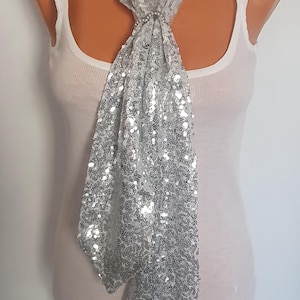 May include: A silver sequin scarf draped over a mannequin. The scarf is made of a shimmering fabric and is adorned with countless small, reflective sequins. The scarf is tied in a knot at the neck, creating a cascading effect.